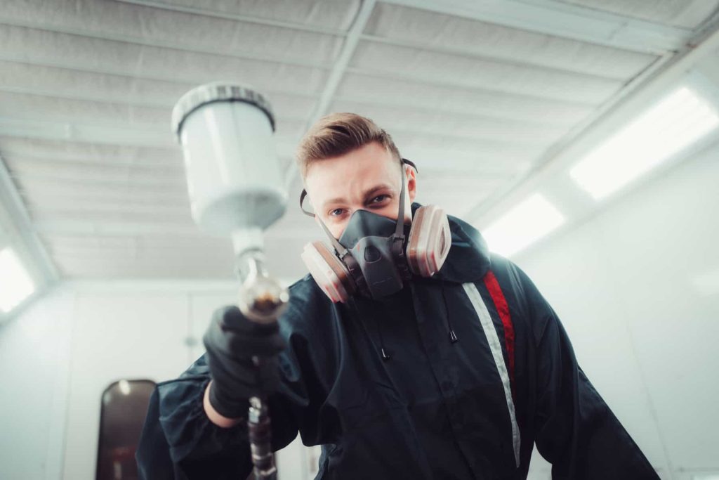 Ein Lackierer mit Atemschutzmaske und Overall hält ein Farbspritzgerät in einer Werkstatt und blickt direkt in die Kamera.