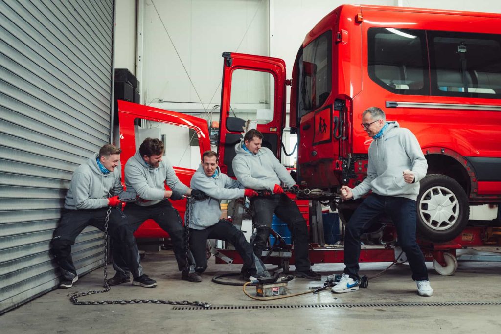 Fünf Mechaniker ziehen in einer Werkstatt eine schwere Kette, die an einen roten Lastwagen gekoppelt ist, und demonstrieren damit Anstrengung und Teamwork.